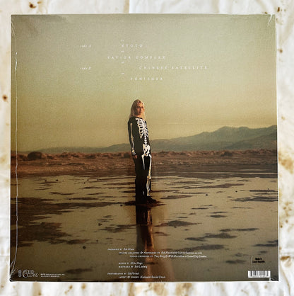 This is the back cover of Phoebe Bridgers & Rob Moose LP record vinyl Copycat Killer. Phoebe is standing on the cover in a skeleton outfit standing straight looking into the camera. She is standing in a muddy area in the middle of a desert 